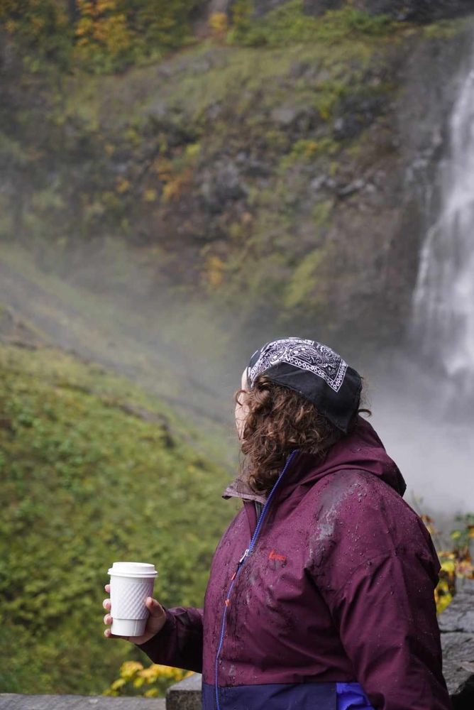 person in a red raincoat and a black bandana holding a hot bev, staring out at the mist 