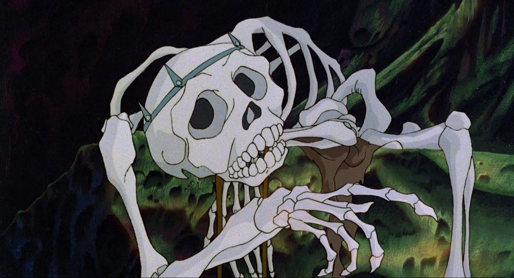 Talking skeleton from The Last Unicorn, slumped on a rocky outcropping of a cave wall, looking very, very drunk and behaving like a desperate addict as he gestures toward something with one long-fingered hand. He wears the remnants of a pewter-colored crown comprised of a thin band topped with 4-5 thin, gemmed spikes.