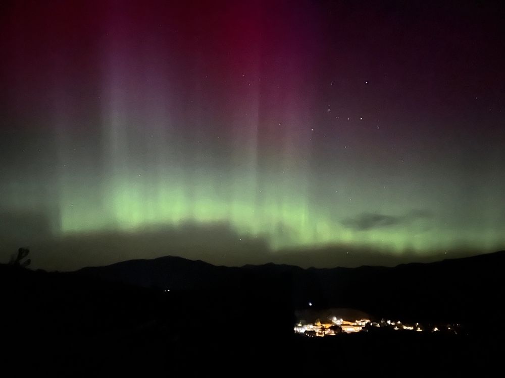 Photo of auroras with purple pillars and green base above the small cluster of lights that is Mammoth, WY.