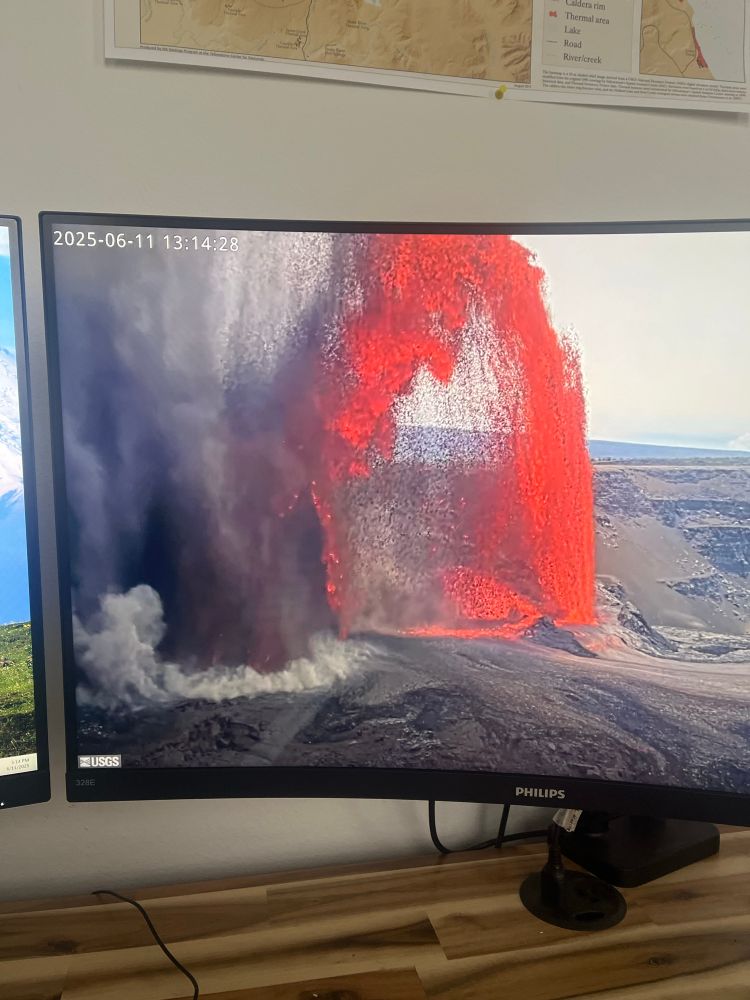Photo of a computer monitor showing an impressively tall lava fountain at Kīlauea which arcs over so completely that it looks like an arch.