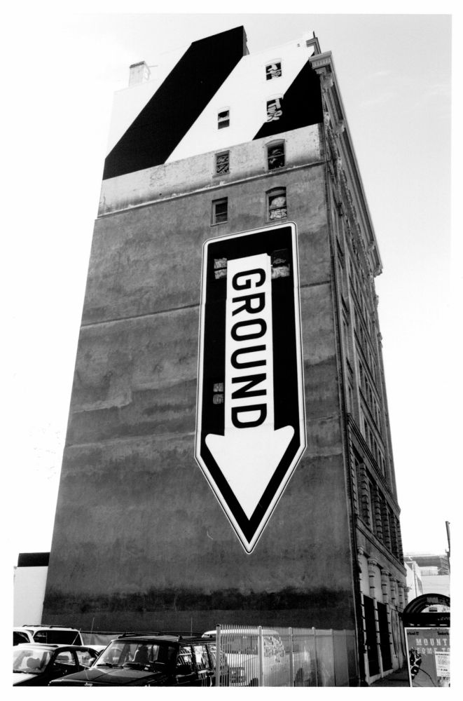 The same classic San Francisco building, showing a mural painted on a different side. The mural is stylized as a one-way traffic sign, pointed vertically downward with the word “GROUND”.