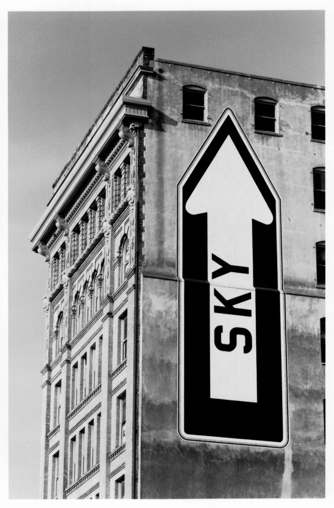 A classic San Francisco building with a mural painted on one side. The mural is stylized as a one-way traffic sign, pointed vertically upward with the word “SKY”.