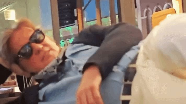  gif of Val Kilmer circa the late 20teens/early 2020s. He's casually lying down on a table or counter of some sort with his head propped on his hand. He's wearing sunglasses, a blue button down shirt, light colored pants, a jacket, and a bandana, as he often did later in life to cover his trach. As the camera pans to his face, he laughs/grins