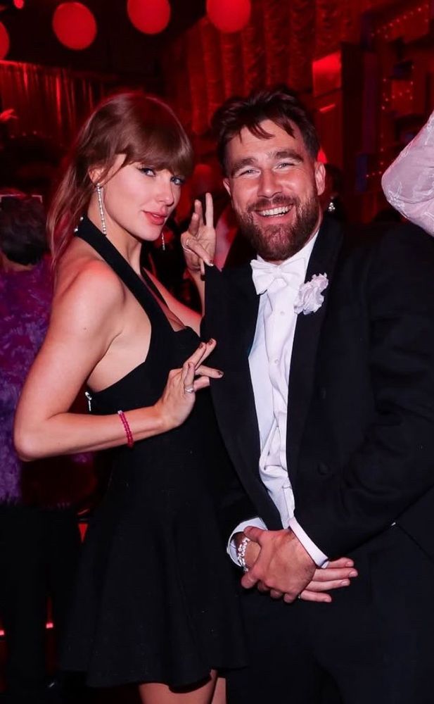 Taylor swift holding the edges of Travis Kelce’s coat jacket. They are both wearing friendship bracelets. Taylor Swift is wearing a black mini dress with a halter top.