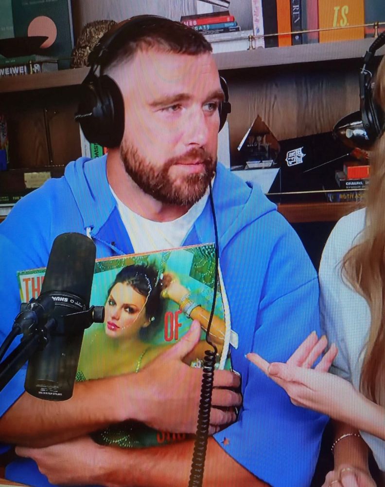 Travis Kelce on the new Heights podcast with Taylor Swift. He is holding the album the life of a showgirl. He is wearing a over the ear headset and wearing a blue zip up jacket. You can see Taylor’s hand on the side of the photo, her shoulder and some of her hair. in the background, you can see miscellaneous items on the shelves, such as trophies, books, little Easter egg boxes that Taylor left