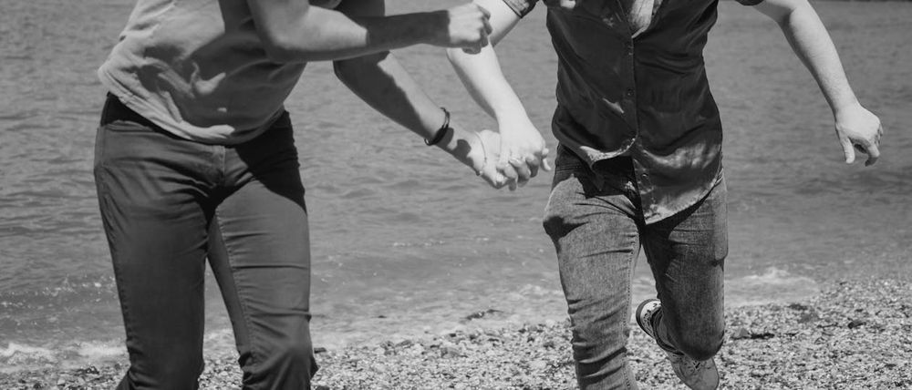A black and white photograph of the torsos and legs of two men are running toward the camera. their faces are not seen, they are holding hands. There is a body of water behind them
