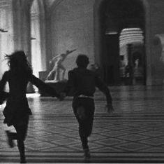 Two people, a woman and a man are running through an empty hallway with a tiled flooring. They are holding hands and the man is running in front of the woman. The photo is black and white, grainy and blurry. A statue of a man pointing at the sky can be seen in the background. 