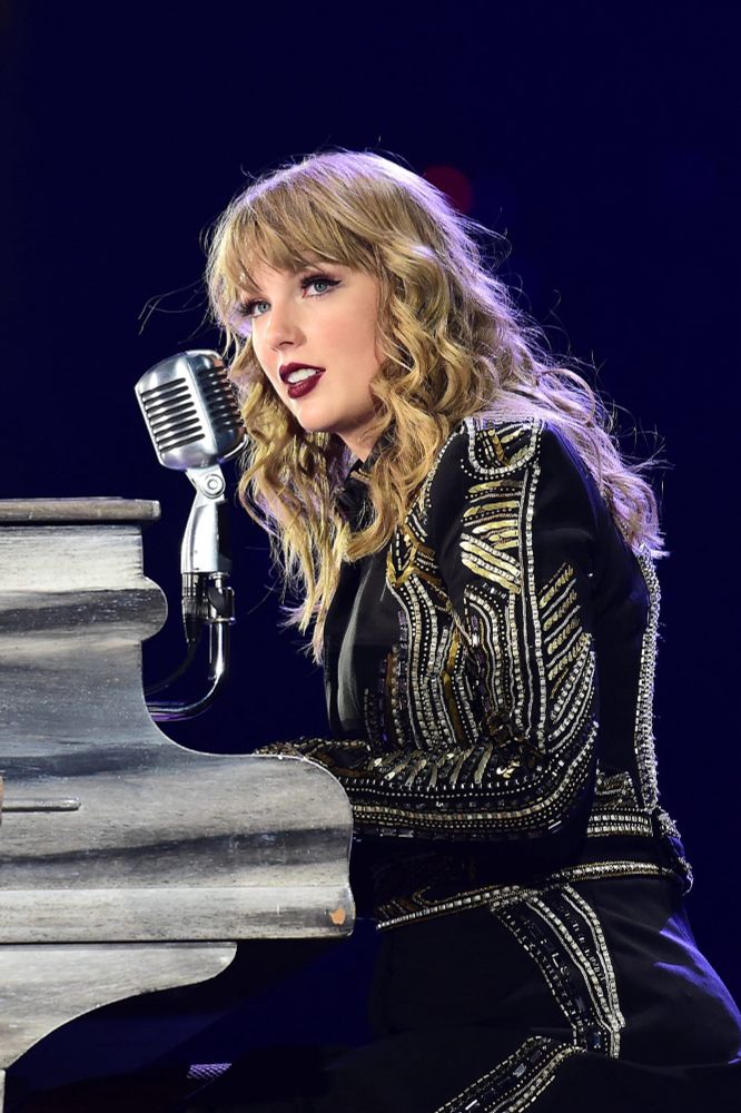 Taylor Swift sits at her piano during the reputation tour, she is wearing a black jacket with gold and silver embellishments 