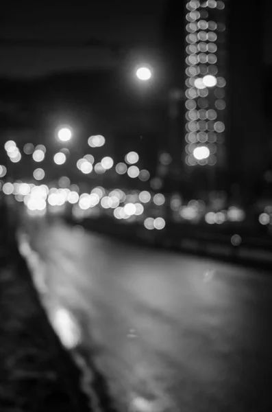 A black and white picture of a darkened street, blurry streetlights and headlights are coming from the left side of the street. A tall building can be seen in the background 