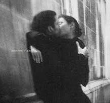 An old style black and white grainy photograph of two people, a woman and a man, embracing in a kiss. There are panes of windows to the right of the photo  