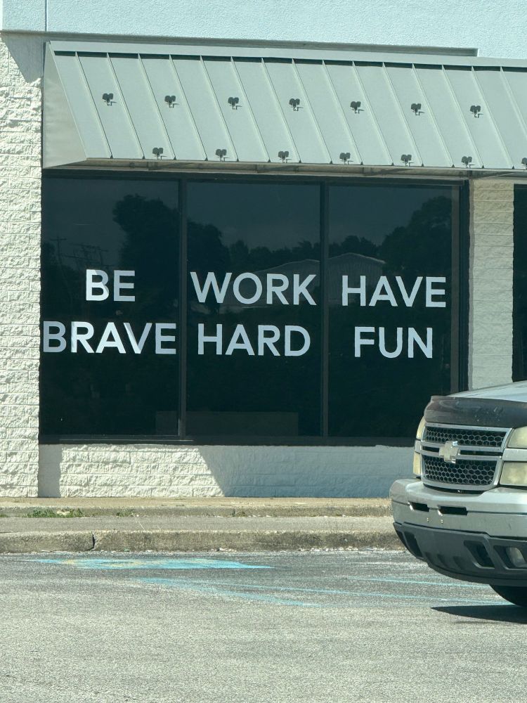 Store window with adhesive letters that say “BE WORK HAVE BRAVE HARD FUN” or Be Brave. Work Hard. Have fun.