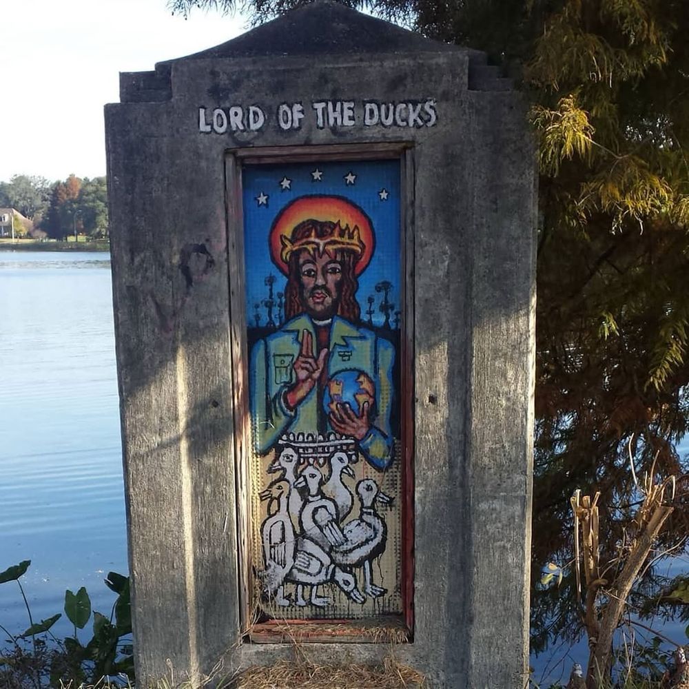 Picture of street art from Baton Rouge, Louisiana. At the top it says “Lord of the Ducks” and beneath depicts Jesus with dark skin and hair wearing a crown of thorns with a hallow behind his head. He holds  globe in one hand and the other is in a posture of blessing. Below him are 6 ducks 