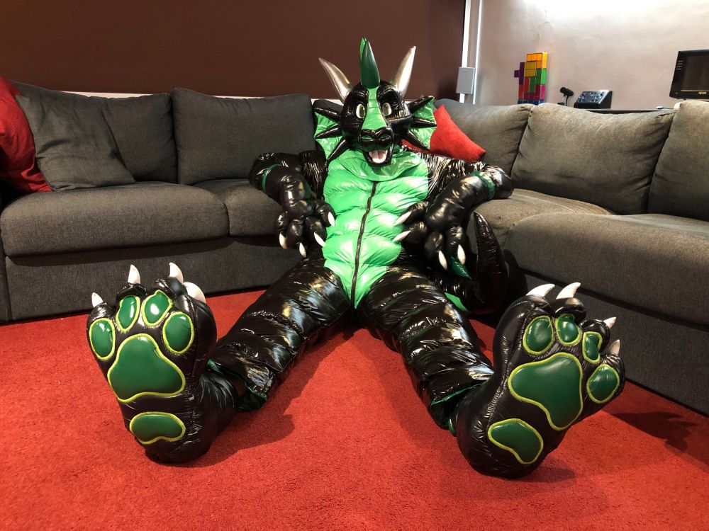 Zorr, the shiny nylon dragon, made of black and green glossy PU coated nylon.  Sat on the floor, stretching out to show off his big soft feetpaws.