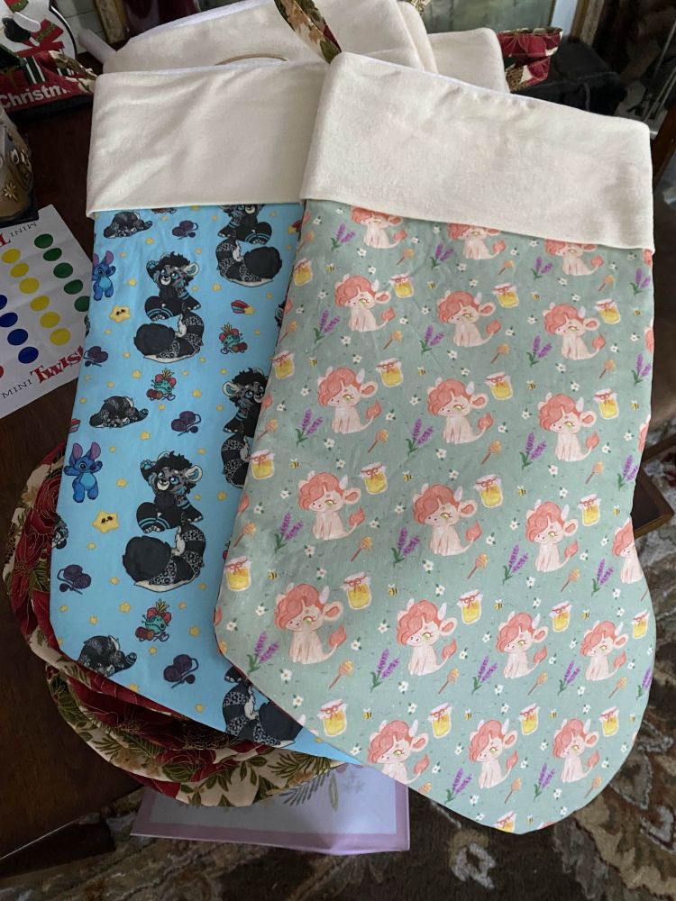 2 stocking are displayed. The first has a custom print of Puck’s sona, a highland cow on a light green background with bees, honey jars, lavender and daisys scattered around. The pattern is repeating. 
Art done by Alexbeeeza on X/twitter.

The second stocking has a custom print of Rikkus sona, a red panda/snow leopard on a blue background with little stars, stitch and scrump plushies and yarn skeins scattered around. The pattern is repeating. 
Art by Honeypuckle.