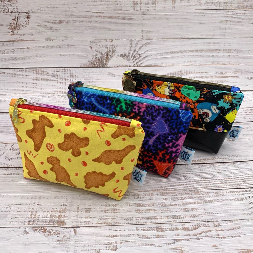 3 snack bags lined up diagonally. First has a dino nugget print on yellow with red swirls. Middle one is a rainbow mushroom print. Last has a Pokémon print with a vinyl bottom.