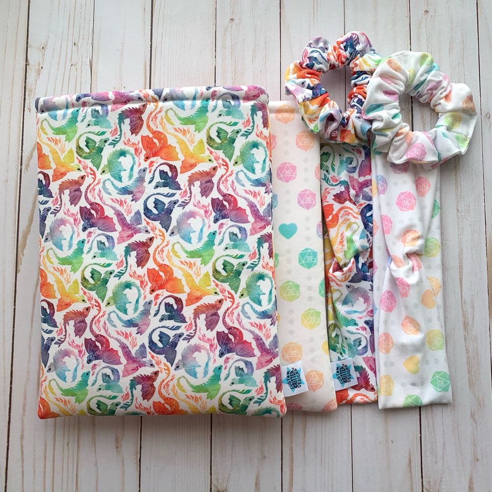 2 booksleeves are stacked on top of each other with 2 headbands to the right of them. The top Booksleeve has a rainbow dragon print and the one below is rainbow d20s and hearts. The headbands both have matching prints.