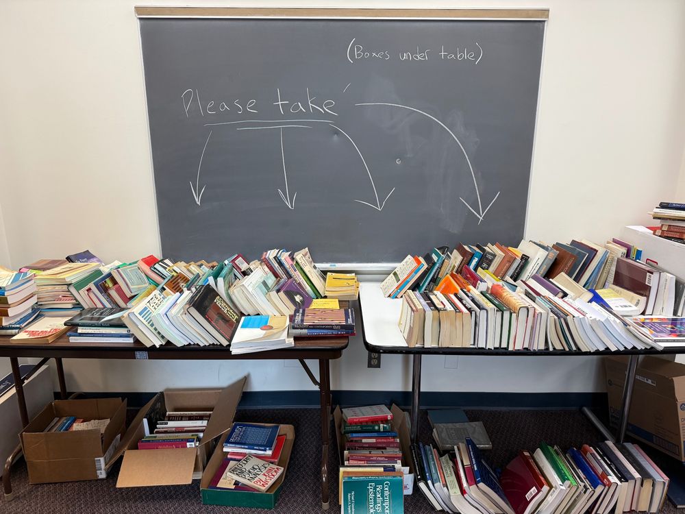 A large pile of books underneath a chalkboard with the words “please take” written above them and arrows pointing toward the book pile. 
