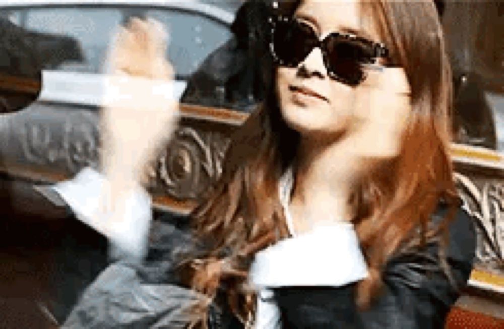 a woman wearing sunglasses is sitting in a car and waving her hand .