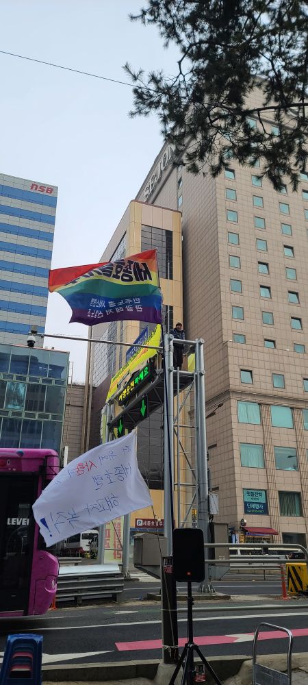 道路の施設物の上で人がが下にあるカメラの方を見ている。道路と人道の中には韓国語で「世宗ホテル支部 民主労総サービス連盟観光レジャー産業労組」と書いているレインボー旗と韓国語で「その上に人がいる 世宗ホテル解雇者復職！」と書いている手旗が翻っている。また、人の後には世宗ホテルが見えている。