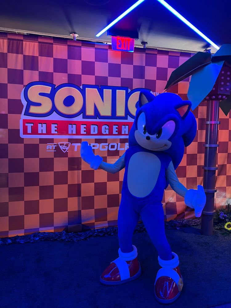 Sonic the Hedgehog mascot standing in front of a “Sonic the Hedgehog at Top Golf” checkered sign 