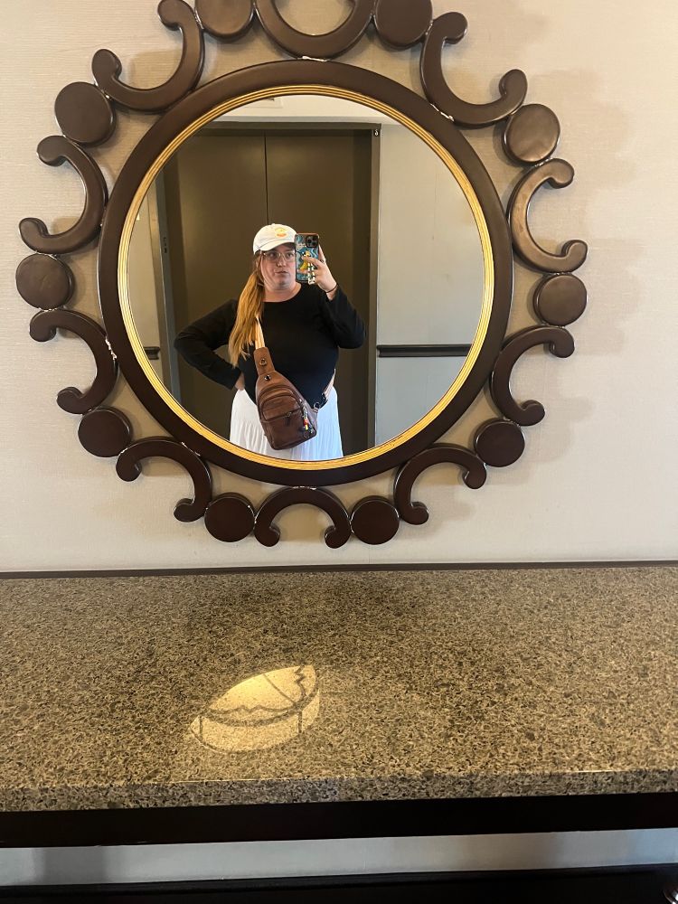 Sydney in a blue hat, long sleeve shirt, and white skirt taking a mirror selfie 