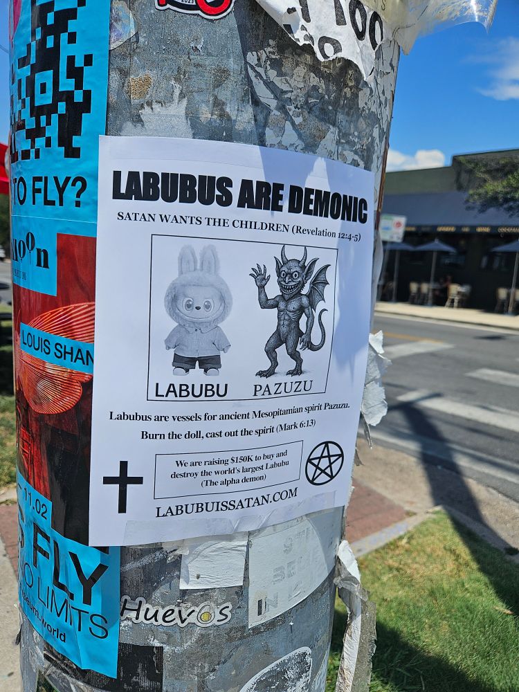 Strange flier taped up on a pole. It has a picture of a Labubu next to a picture of a strange demon apparently called a Pazuzu. In one bottom corner there is a cross and in the other there is a pentagram. 

The text is as follows:

LABUBUS ARE DEMONIC
SATAN WANTS THE CHILDREN (Revelation 12:4-5)

Labubus are vessels for ancient Mesopitamian spirit Pazwa. Burn the doll cast out the spirit (Mark 6:13)
We are raising $150K to buy and destroy the worId's largest Labubu (The alpha demon) LABUBUISSATAN.COM