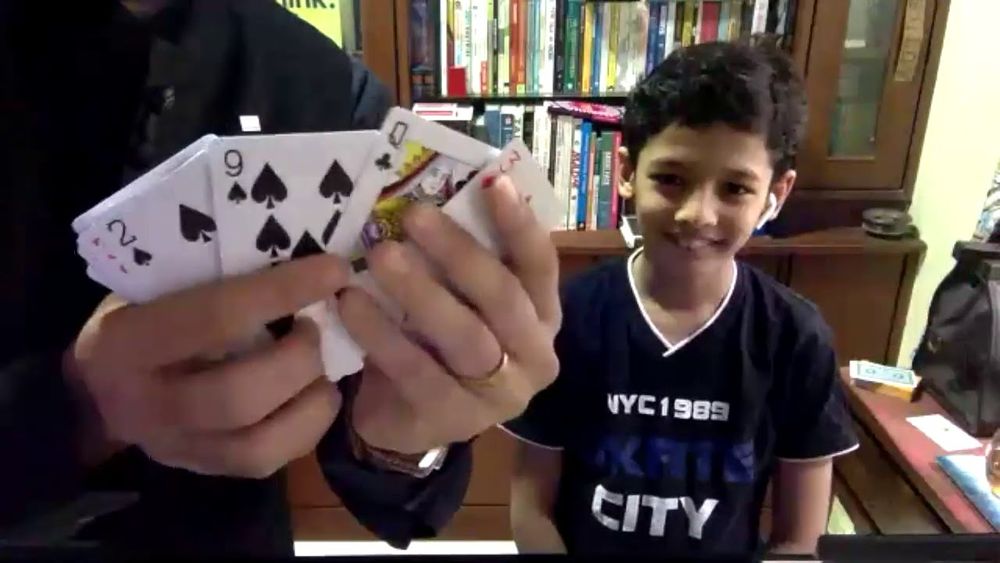 The photo shows a person (me) holds a fan of playing cards showing the 2, 9, and Queen of Spades, along with another card partially visible. In the background, a smiling boy wearing a dark T-shirt with ‘NYC1989 CITY’ printed on it is sitting in front of a bookshelf filled with books and a wooden cabinet. (Alt text generated by GPT.)