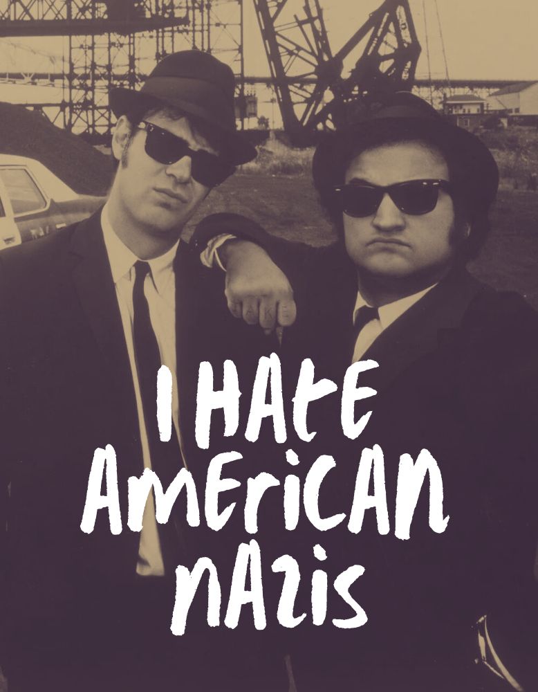 Dan Aykroyd and John Belushi as the Blues Brothers. Dan as Elwood Blues is on the left. He is standing with his head tilted toward the center of the frame; looking directly at the camera. John as Jake Blues is on the right. He is standing with his elbow resting on Elwood’s shoulder. His hand is in a fist, knuckles towards the camera. He is also looking at the camera; his right eyebrow is raised. Both men are dressed in their signature black suit, with narrow tie, black hat, and sunglasses. They are standing in a disused factory yard. Their iconic blues mobile (former black and white police car) is about 10 feet behind Elwood on the left side of the photo. The photo has been edited to a sepia tone. In bright white, centered on the lower third of the photo, are the words “I hate American Nazis”. The font looks like scribbled graffiti.