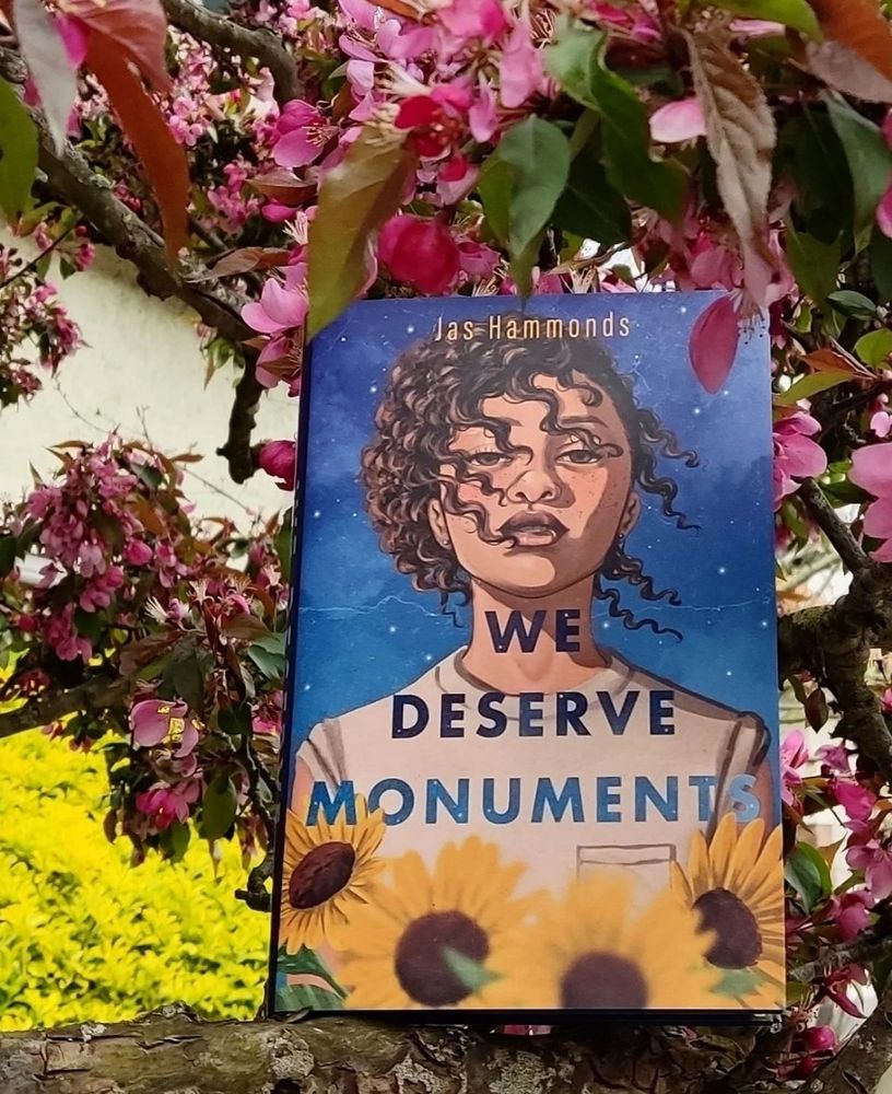 The book We Deserve Monuments by Jas Hammonds on a tree branch with pink flowers behind it.