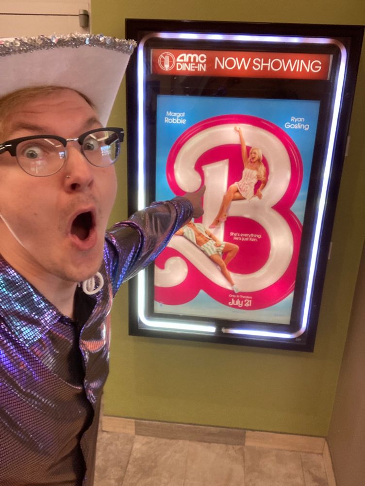 Photo is of me staring at the camera with my mouth agape pointing at the Barbie movie poster in my Barbie movie outfit consisting of a white sequined cowboy hat and an iridescent purple button up shirt with a disco ball pattern.