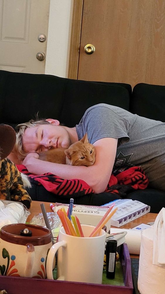 Human is laying down on a futon. Head is resting on Rajah the orange tabby’s ample bosom with their arm encircling him. They are snuggling up to one another.
