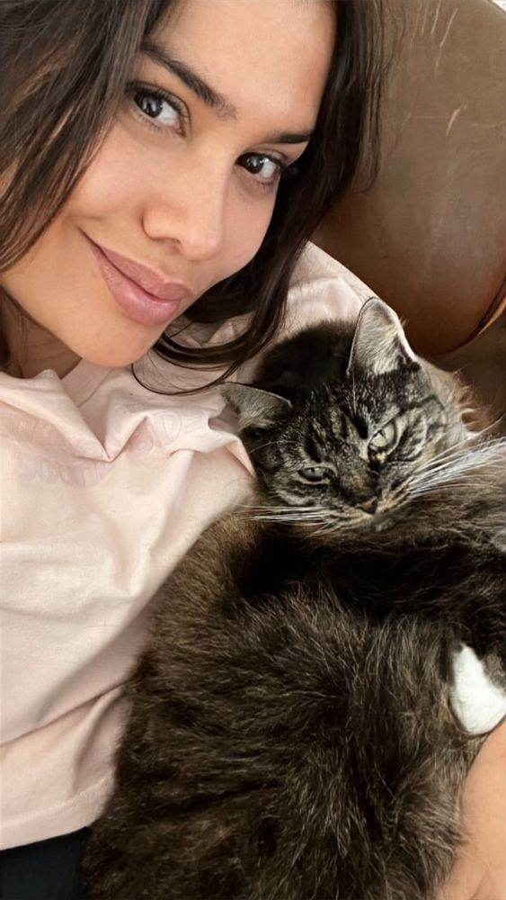 Em close, Leila de camiseta rosa calro numa poltrona de couro segurando a sua gatinha Paçoca que é tigrada e cinza, esparramada com a cabeça em seu ombro. A gatinha encara a câmera também. Não dá pra ouvir mas ela estava ronronando. 