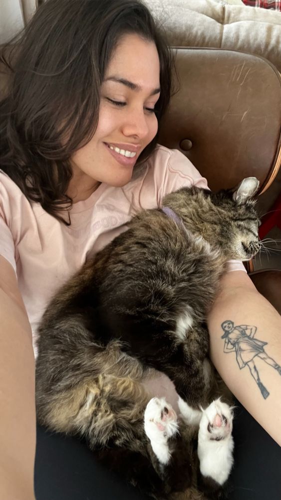 Leila numa poltrona de couro segurando a sua gatinha Paçoca que parece totalmente entregue e manhosa em seu colo. Leila olha sorridente para a gatinha felpuda e tigrada em cinza, que por sua vez dorme esparramada com a cabeça em seu ombro, enquanto fica de barriguinha pra cima. A imagem é vertical e pega da cabeça de Leila até a coxa. 