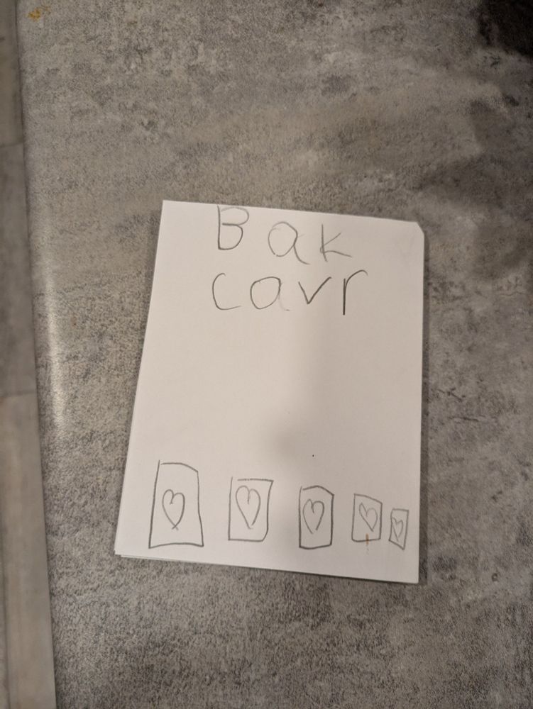 Small piece of paper. A child's handwring. "Bak covr", with an eraser mark showing that the o used to be an a. Hearts. 