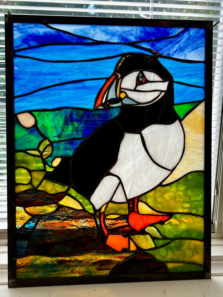 An Atlantic Puffin executed in stained glass. The bird has a black body with white belly, black head, white face, orange and gray eyes, orange legs and feet, multi colored bill (orange, grey and yellow) with yellow at its base. It is standing facing right but with head turned to the left. It is on a lower background of green, yellow, and brown, and an upper background of the blue sea and cloudy sky. 