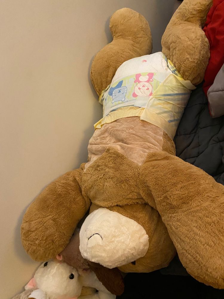 Overhead view of large, about four foot long simplistic dog plush in a tykables animooz diaper, laying belly up with head towards the camera, on a bed with a weighted blanket and comforter. A smaller white weasel plush is also visible in the bottom left corner.   