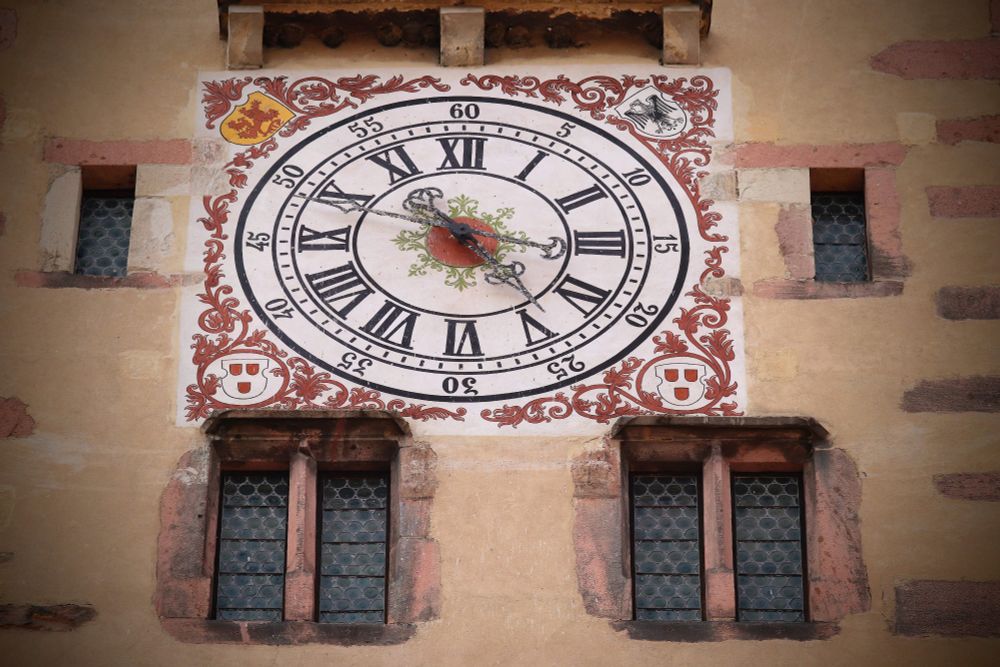 19 century clock on the wall of a medieval tower. Pink floral patterns on white enamel. The numbers of the clock are dark blue. There are coats of arms in each of the four corners