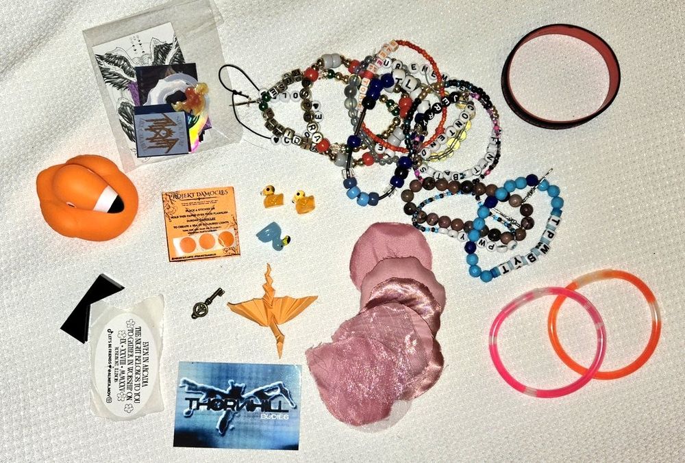 Sleep Token and Thornhill trinkets from a concert spread out, including bead bracelets, stickers, origami, flamingo figures, squeaky flamingo duck, glow sticks, and petals from stage/pit