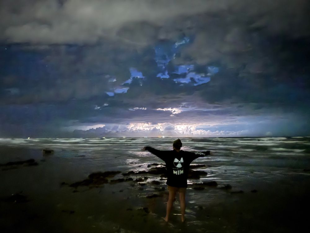 Girl facing the beach at night wearing motionless in white hoodie where the words are glowing, she is standing on the beach with arms open looking out at the as a lightning storm is in the distance.