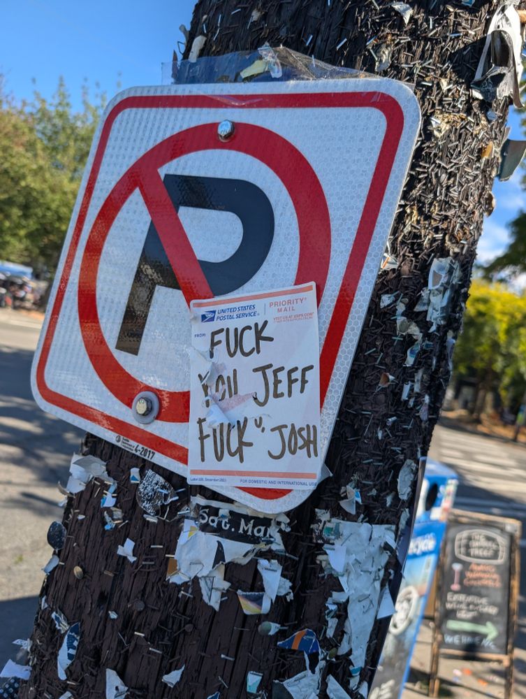 A no parking sign with a USPS label on it, upon which someone has written FUCK YOU JEFF. FUCK U JOSH 