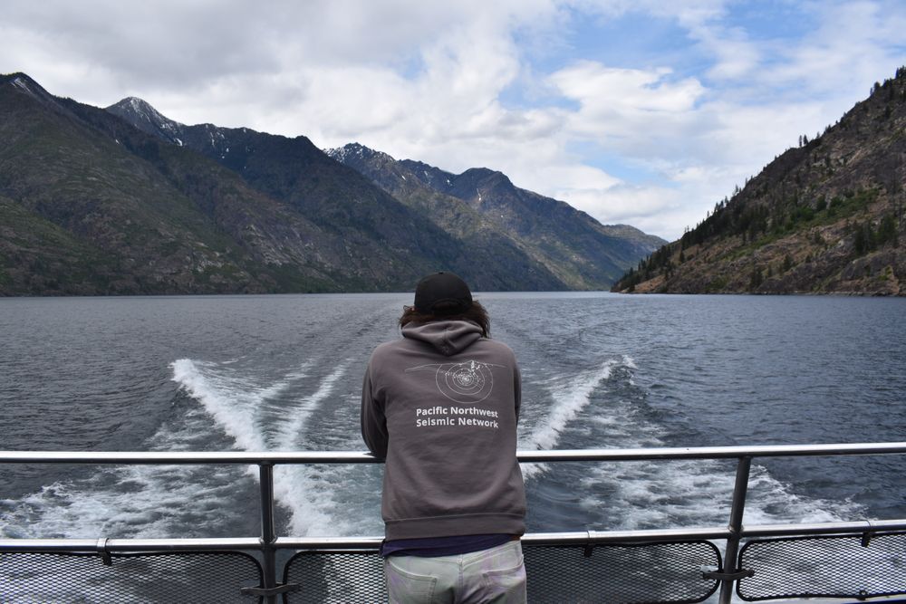 PNSN Field Technician in grey sweatshirt standing along metal ferry railing with mountains in background on both sides of Lake Chelan.