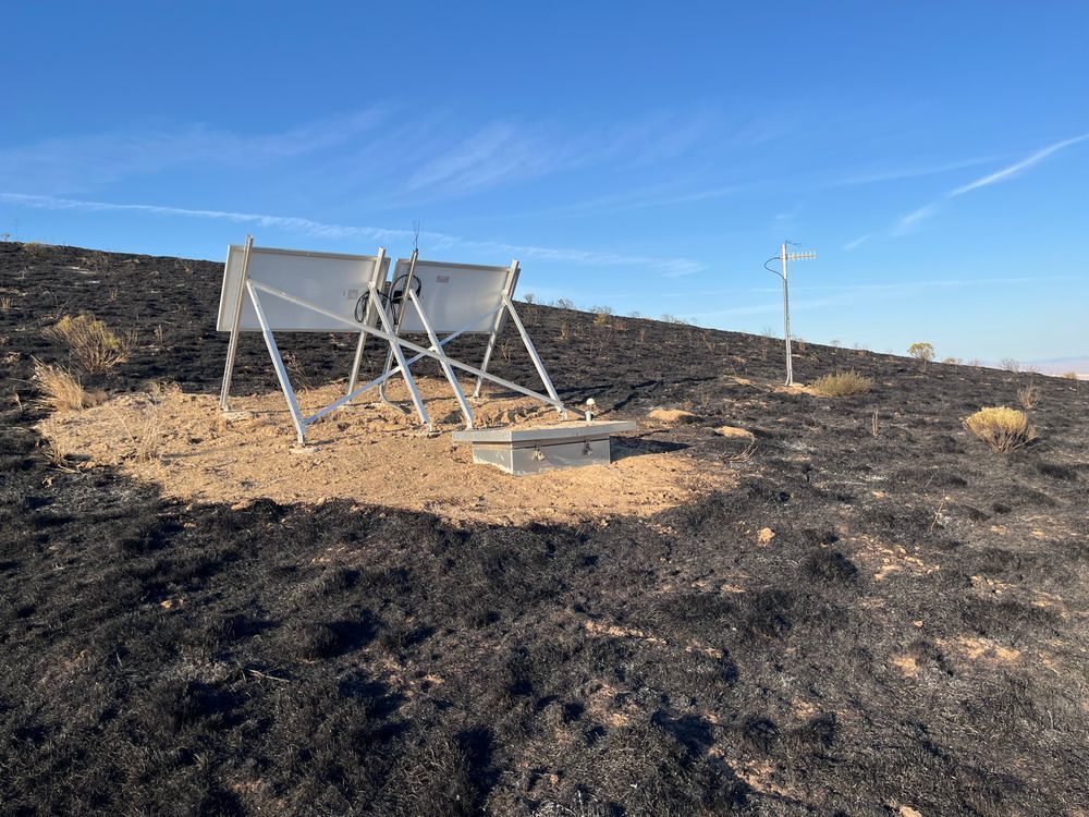 Two solar panels from behind on a dark soil hill. The sky is clear and there is a short antenna sticking out of the ground near by. 