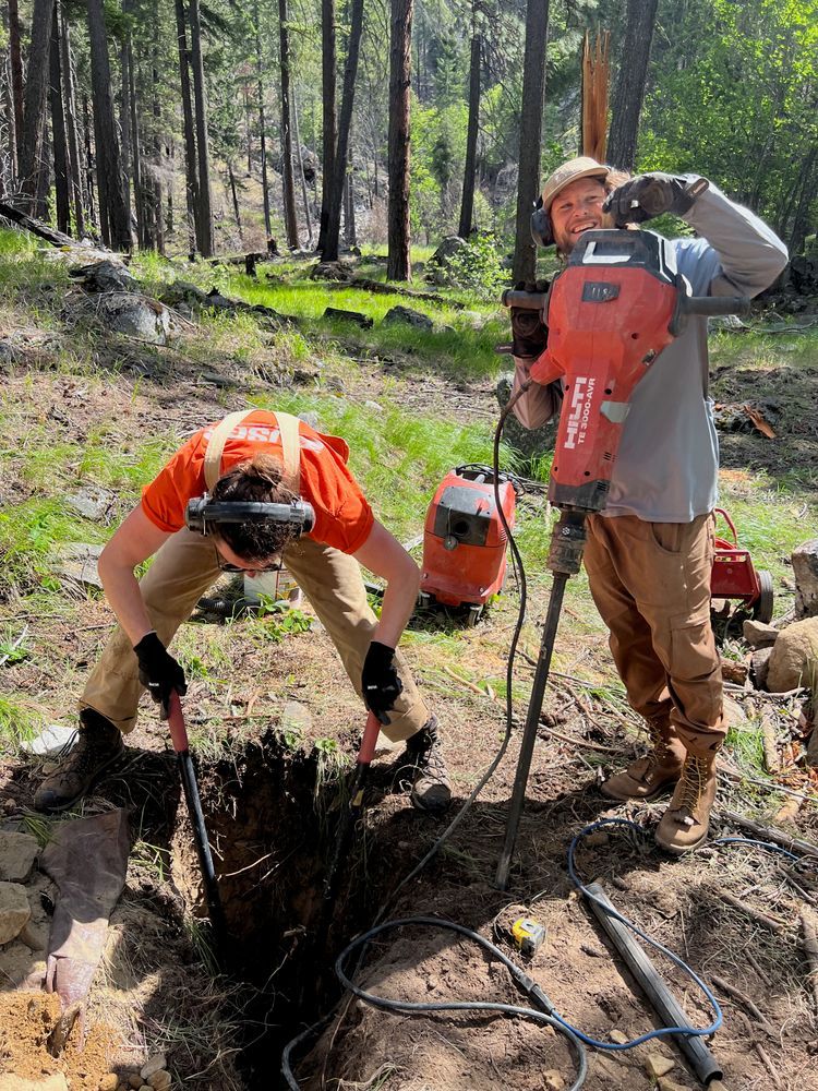 Field Technicians digging in vault hole in the ground with another field tech holding large red jackhammer with 3ft bit attached. Forest in background.
