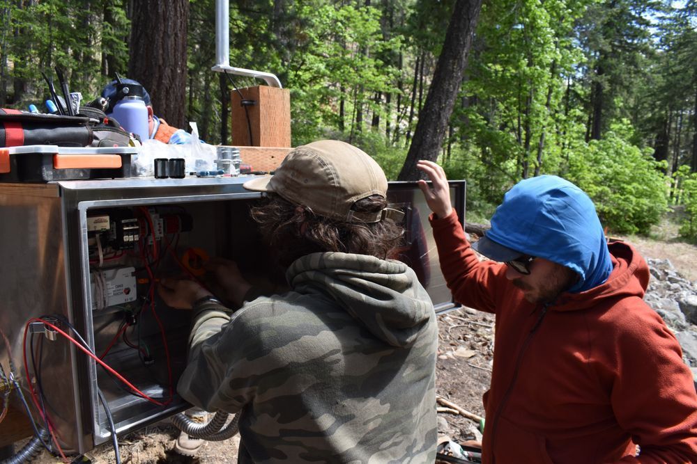 Field Technicians working on seismic station electronics box. Forest in background.