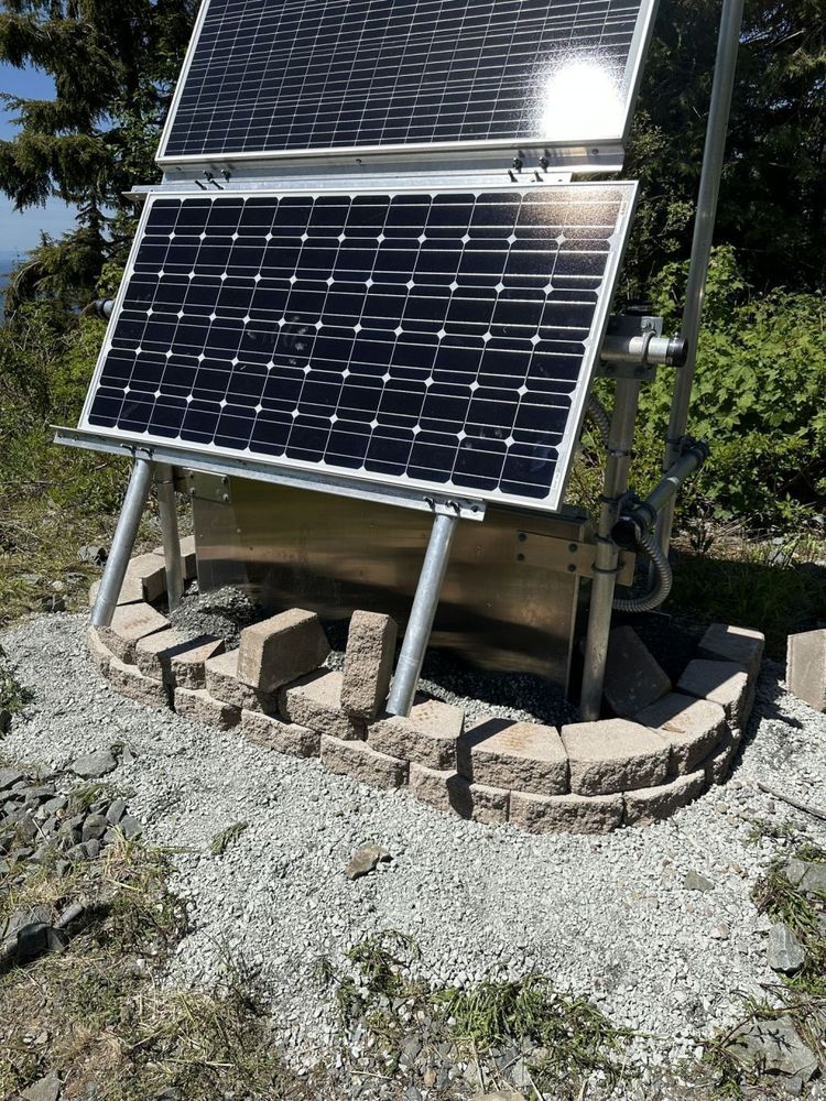 Paving bricks and gravel being laid along the southern side of seismic swing set structure. Black solar panels attached to metal box. Forest in background.