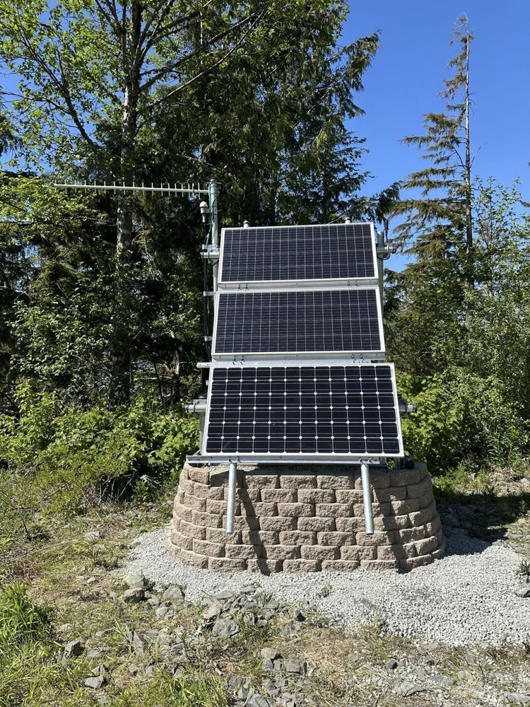 Completed paving brick wall and gravel emplacement around seismic structure. Black solar panels and long horizontal antenna at top. Forest in background. 