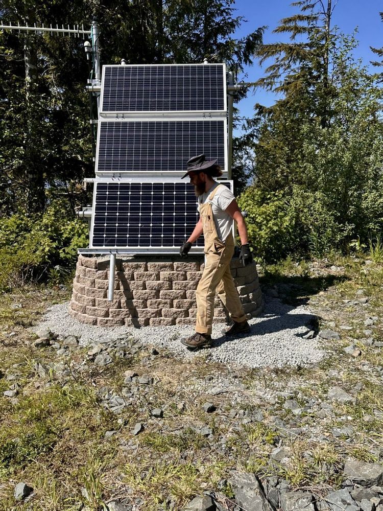 Field Technician walking in front of paver wall and seismic structure black solar panels. Forest in background. 