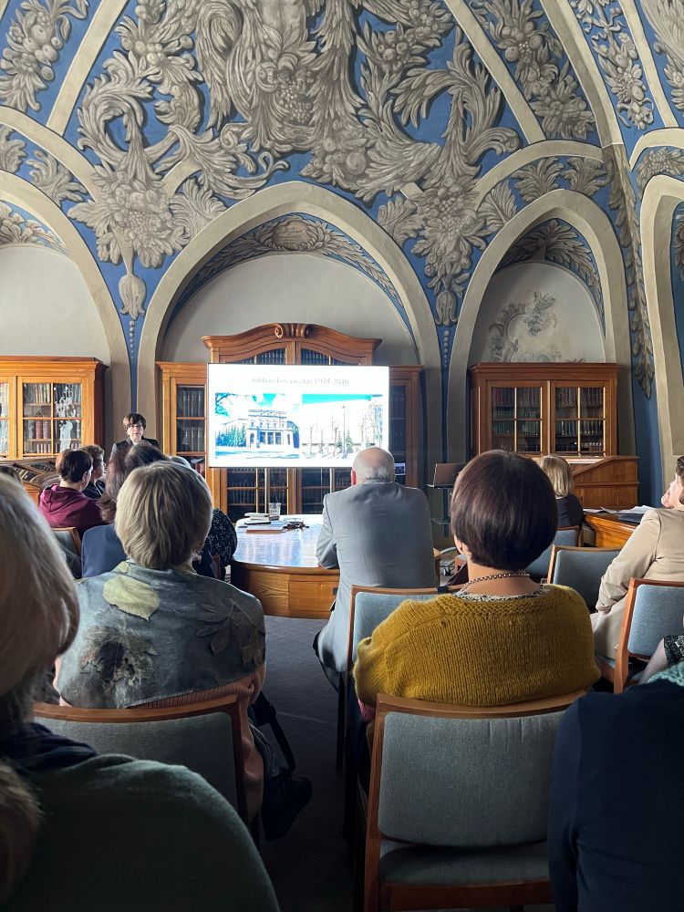 A group of people sits in a historic Vilnius Universtity library room with an ornate vaulted ceiling decorated in blue and gray floral patterns. 
