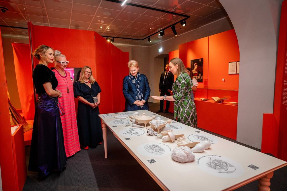 In a vivid orange exhibition space, former President Dalia Grybauskaitė listens as Dr. Milda Kvizikevičiūtė (in a green patterned dress), one of the curators, presents a display table featuring ceramic reconstructions of historical dishes, biscuit moulds, vegetables, and a frying pan. The table is illustrated with recipes and foods once enjoyed by Renaissance queens Barbara Radziwiłł and Catherine Jagiellon. Nearby stand co-curators Dr. Vaida Ragėnaitė (in a violet dress) and Julija Janus (in pink stripes), along with Rūta Kačkutė (in a dark blue dress), general director of the National Museum of Lithuania. Exhibition cases with rare books and portraits are visible in the background.