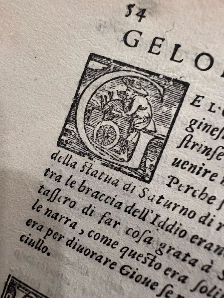 Engraved initial “G” from an early printed book, featuring a seated or crouching figure wearing a hat, pushing a cart filled with plants.